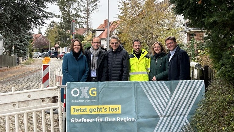 Gruppe von sechs Personen steht vor einem Banner mit der Aufschrift „Jetzt geht's los! Glasfaser für Ihre Region“ in einer Straße, die mit Baumreihe und Wohnhäusern gesäumt ist in Berlin Tempelhof-Schöneberg